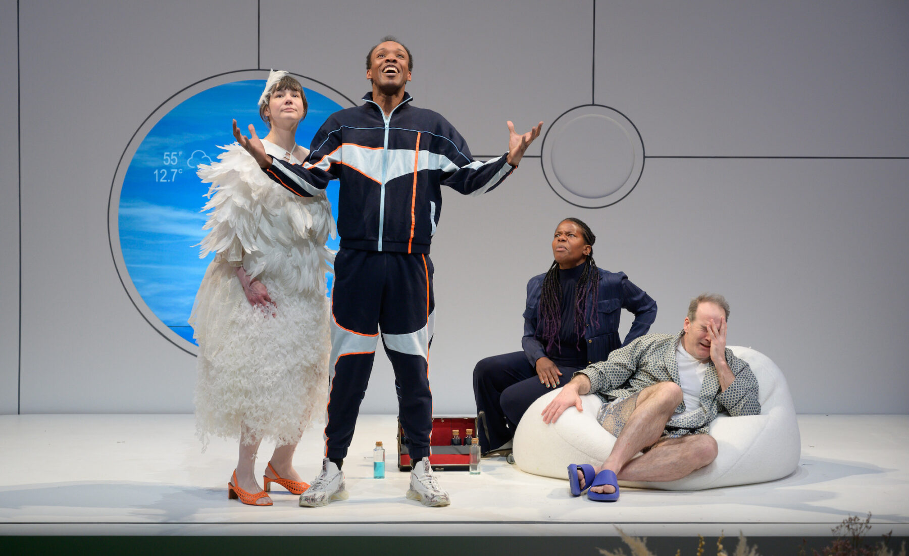 Brandon J. Pierce stands with his arms out wide looking joyously up into the sky. He is in a sweat suit in various shades of blue and an orange stripe. Sarah Gliko looks on with him standing just behind his right side, still in her white feathers, now in orange kitten heels. To Peirce’s left side, his parents sit on a bean bag chair, looking surprised. Jennifer Kidwell is in an all-navy business casual suit and Steven Richard is in a light blue robe, underwear and slippers.